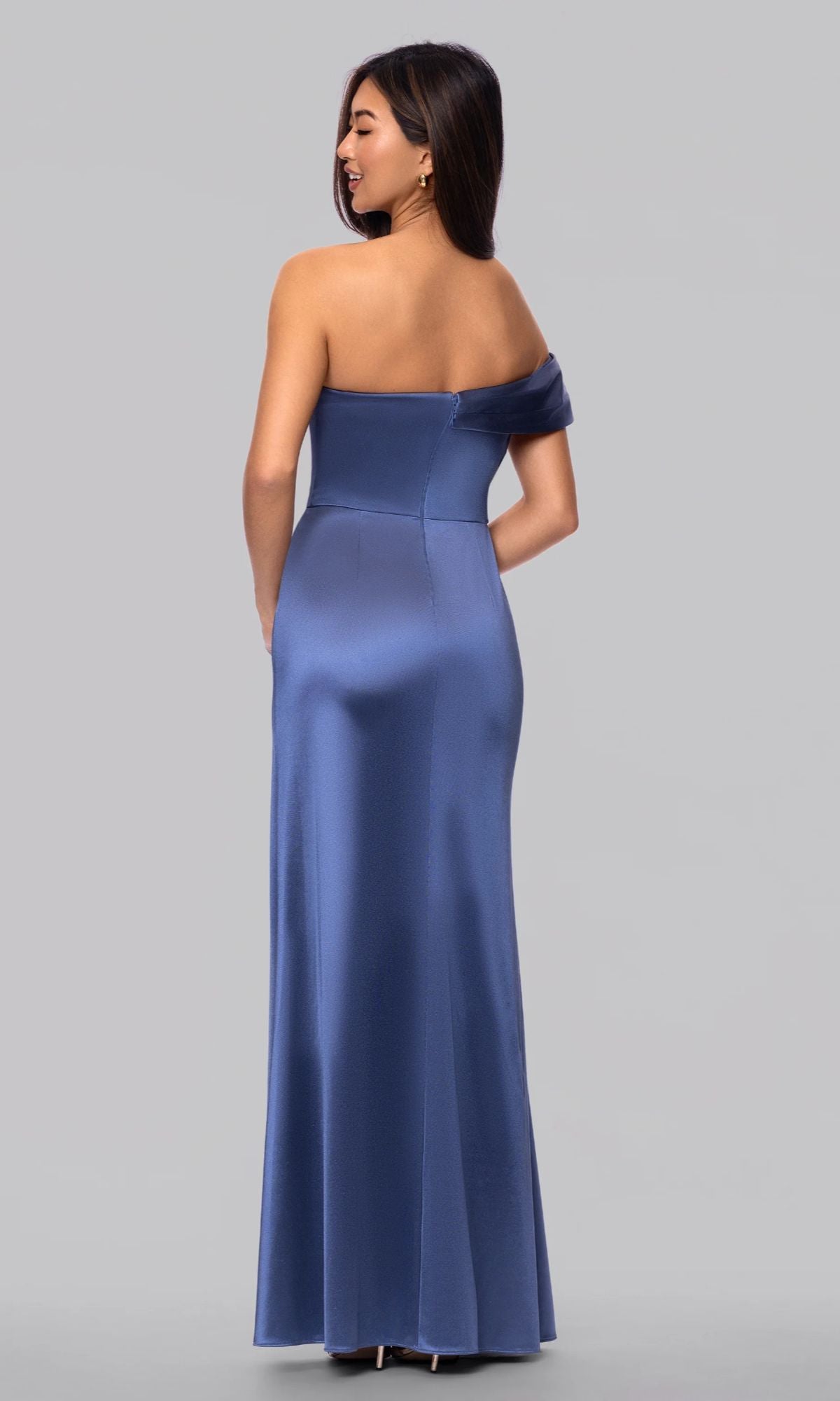 A woman faces away, wearing the Blondie Nites 6164BN long satin off-the-shoulder blue prom dress, standing against a plain gray background.